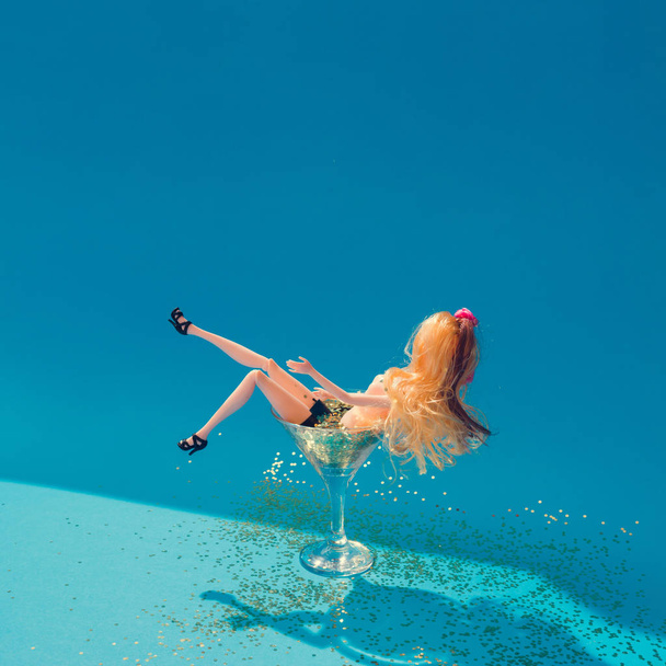 Poupée se baignant dans un verre de martini plein de paillettes dorées sur fond bleu, concept d'été de beauté minimale créative
  - Photo, image