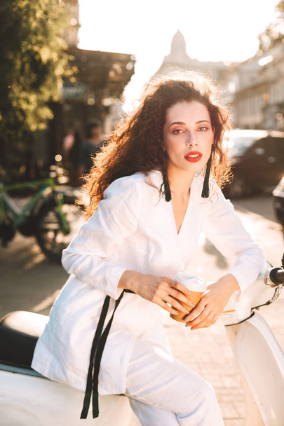 Jovem bela senhora com cabelos encaracolados escuros em traje branco sentado em branco ciclomotor com xícara de café para ir e sonhadoramente olhando na câmera na rua
 - Foto, Imagem