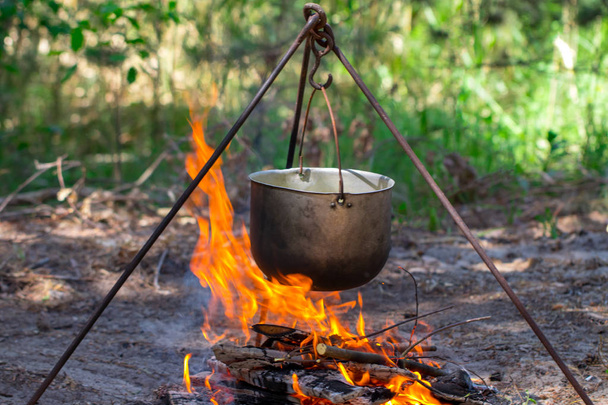 Τουριστική κατσαρόλα κρέμεται πάνω από τη φωτιά σε ένα τρίποδο. Μαγείρεμα στην καμπάνια. - Φωτογραφία, εικόνα