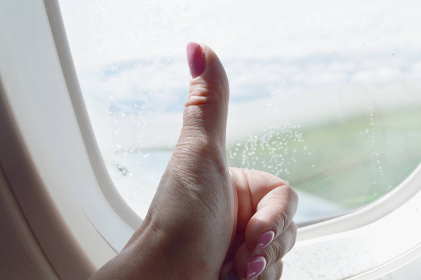Το γυναικείο χέρι δείχνει τον αντίχειρα επάνω, κάθεται στο αεροπλάνο στο παράθυρο. Η πτήση - Φωτογραφία, εικόνα