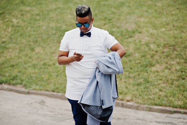 Stylish arabian man with jacket, sunglasses and bow tie posed outdoor and looking on mobile phone. Arab model boy. - Photo, image