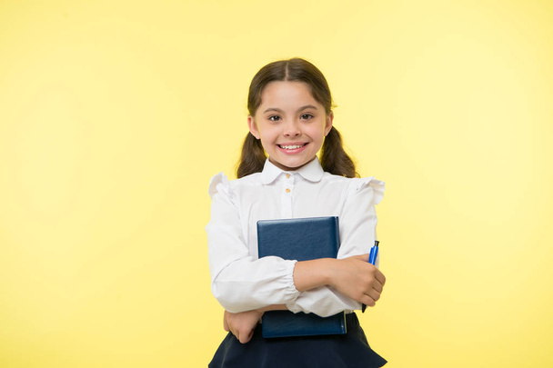 I am ready for school. Child school uniform smart kid done with homework. Girl cute happy face yellow background. Child girl school uniform clothes holds book and pen. Back to school concept - Foto, afbeelding