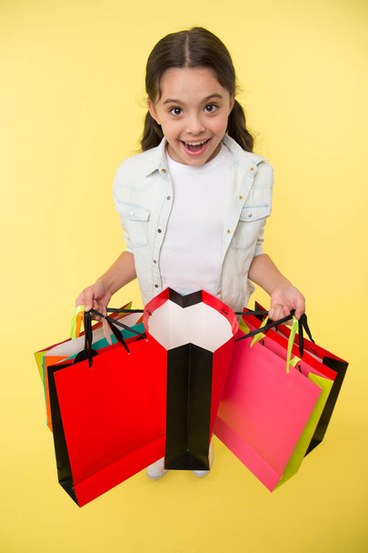 Check it out. Child cute fashionista shopping. Kid girl happy smiling face carries bunch packages yellow background. Girl shopaholic likes shopping on sale season. Kid happy shopping in mall - Valokuva, kuva