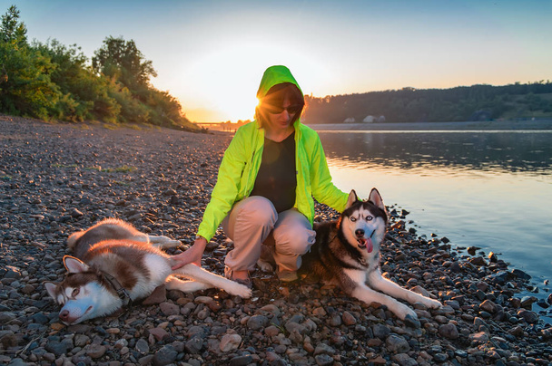 Girl in sunglasses and yellow windbreaker with husky dogs on the river bank in the shine evening sun. Portrait mistress with dogs. - Foto, imagen