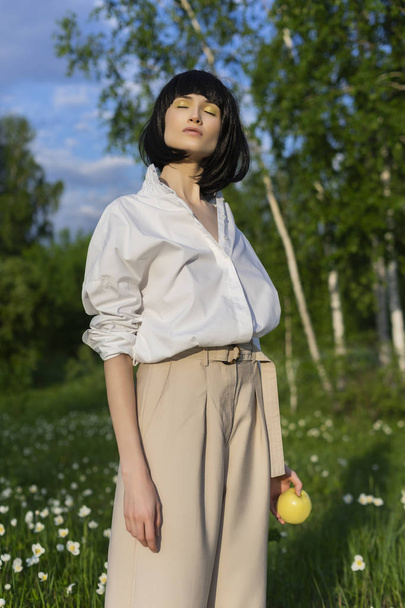 Beautiful stylish girl wearing a trendy white shirt, beige trousers and a black wig holds a green apple in sunlight on a background of a blue cloudy sky. Advertising and fashion design. Copy space. - Fotó, kép