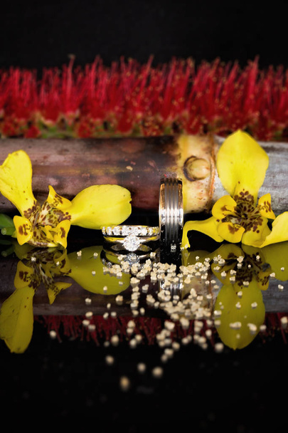 Trouwringen op een glanzend zwarte ondergrond met gele en rode bloemen en suikerkristallen voor hen van suikerriet landbouw - Foto, afbeelding