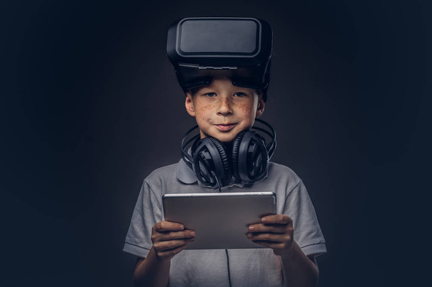 Portrait of a cute little boy dressed in a white t-shirt, with virtual reality glasses and headphones is holding a digital tablet at a studio, looking at a camera, isolated on a dark background - Photo, Image