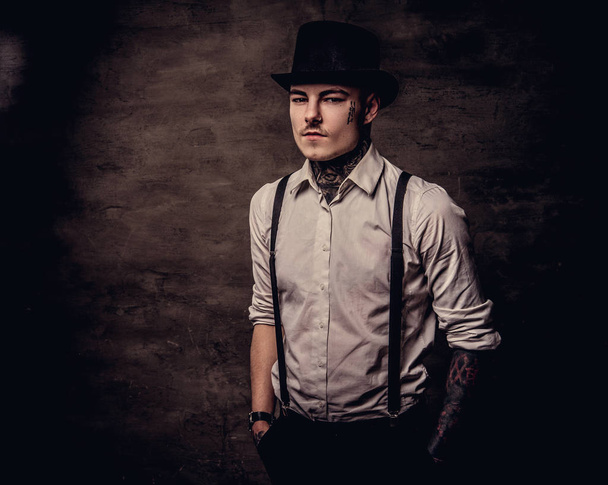 Young old-fashioned tattooed guy wearing a white shirt with suspenders and cylinder hat, looking at a camera. Isolated on a dark background. - Photo, Image