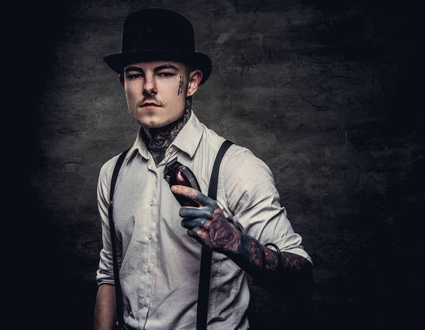 Πορτρέτο του ένα ντεμοντέ τατουάζ κομμωτήριο φορώντας ένα άσπρο πουκάμισο με ζαρτιέρες και κύλινδρο καπέλο, βλέπουν μια φωτογραφική μηχανή, κατέχει ένα χορτοκοπτικό. Απομονωμένη σε ένα σκοτεινό φόντο με υφή. - Φωτογραφία, εικόνα