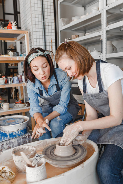 Two girls friends smiling and talking while working on potters wheel making clay handmade craft in pottery workshop, friendship and guidance concept - Photo, Image