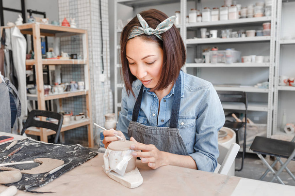 Uma artista mulher no avental de trabalho sorri e pinta uma cerâmica de barro entre escovas e outras ferramentas na oficina moderna
 - Foto, Imagem