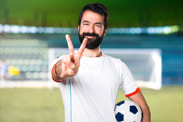 Voetbal speler bezit is van een voetbal bal maken overwinning gebaar op een voetbalveld - Foto, afbeelding