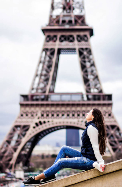 Hermosa joven en jeans y en un suéter blanco, sosteniendo un café en una comida para llevar en el fondo de la Torre Eiffel. Concepto de turista en París
 - Foto, Imagen