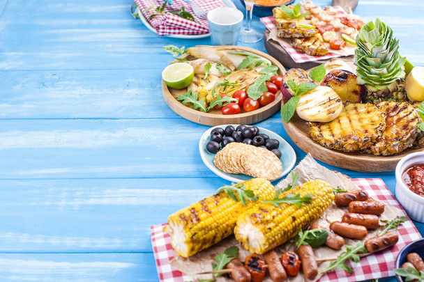 Grilled vegetables, shrimp, fruit on a wooden plate and sausages, juice and salad on a blue background. Summer dinner. Free space for text. Copy space - 写真・画像