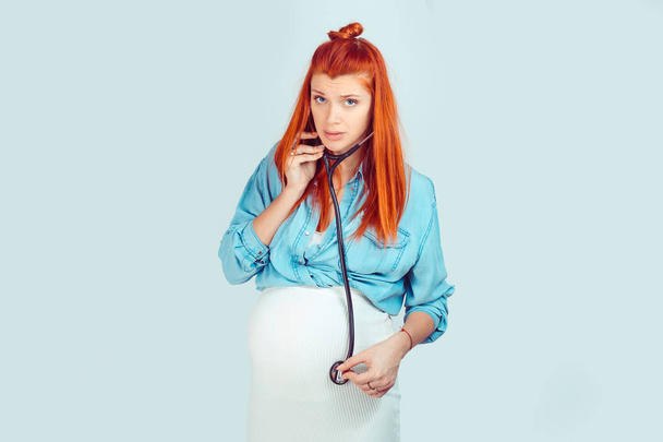 Young ginger pregnant woman using stethoscope and listening to baby with worried face isolated on light blue background. Bad self diagnosis during pregnancy concept - 写真・画像
