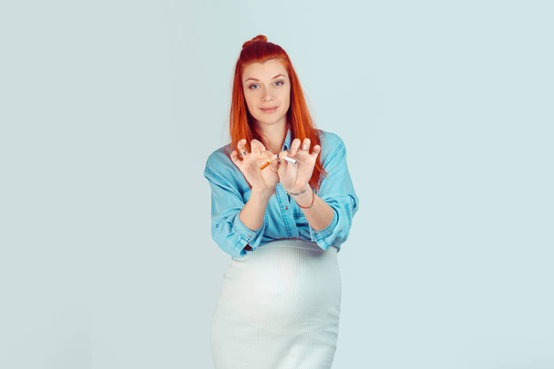 Beautiful young ginger woman in expectancy breaking cigarette with serious expression on face at camera isolated on light blue background. Mixed race model, latin hispanic irish woman - Photo, image