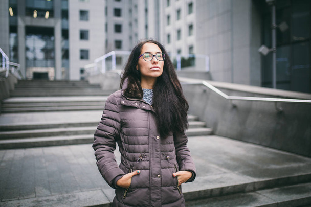 Gyönyörű, fiatal nő európai etnikai hosszú barna hajú, szemüveges, és egy kabátot áll ellen hátteret a business center a csökkenése felhős, felhős Időjárás. - Fotó, kép
