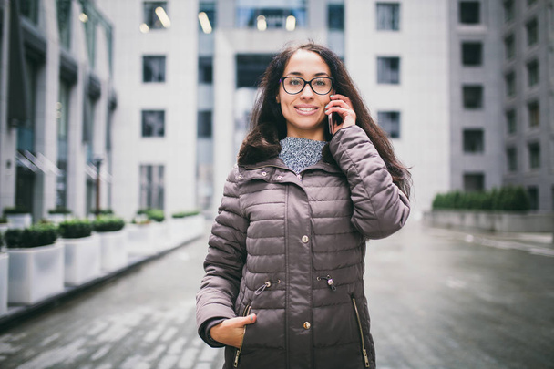 Theme is the business situation. Beautiful young woman of European ethnicity with long brunette hair wearing glasses and coat stands on background of business center and uses phone in hand near ear. - Photo, Image