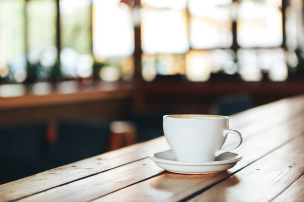 Ζεστό καφέ στο φλυτζάνι λευκό ξύλινο τραπέζι. Λευκό φλιτζάνι του καφέ σε ένα ξύλινο τραπέζι σε μια καφετέρια. Καλημέρα με ένα κεραμικό λευκό φλιτζάνι καφέ σε μια καφετέρια  - Φωτογραφία, εικόνα