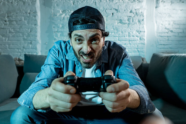 young excited latin man at home sitting in living room on  sofa and  playing video games using remote control joystick with freak intense face expression having fun in gaming addiction concept - Foto, imagen
