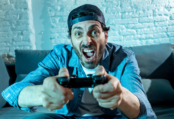young excited latin man at home sitting in living room on  sofa and  playing video games using remote control joystick with freak intense face expression having fun in gaming addiction concept - Photo, image
