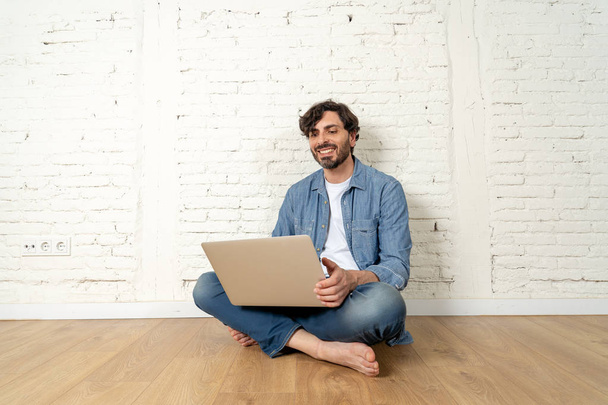 portrait of latin man dressed in T-shirt blue shirt and jeans sitting against white brick wall holding laptop and watching media with happy smile. hipster freelance working from home concept. - Фото, зображення