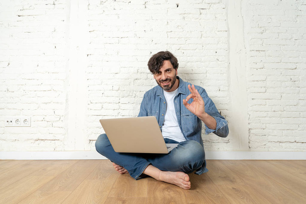 portrait of latin man dressed in T-shirt blue shirt and jeans sitting against white brick wall holding laptop and watching media with happy smile. hipster freelance working from home concept. Man showing ok  up - Fotó, kép