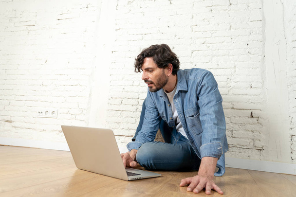 portrait of latin man dressed in T-shirt blue shirt and jeans sitting against white brick wall holding laptop and watching media with happy smile. hipster freelance working from home concept. - Foto, Bild