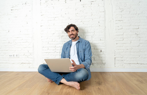 portrait of latin man dressed in T-shirt blue shirt and jeans sitting against white brick wall holding laptop and watching media with happy smile. hipster freelance working from home concept. - Фото, зображення
