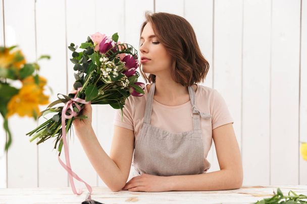 Young cute florist woman standing with flowers in workshop smell it. - Foto, Bild