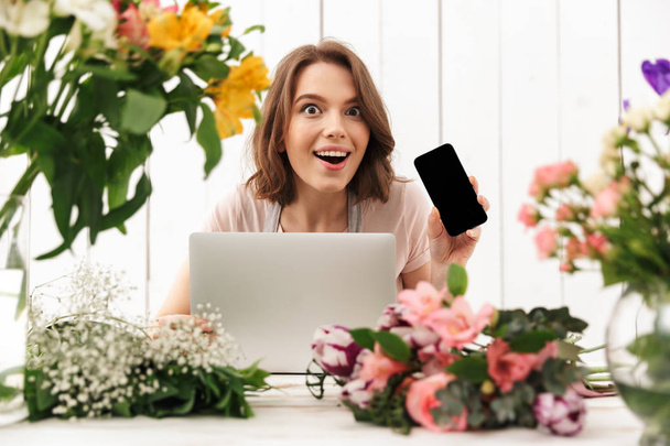 Young cute cheerful florist lady standing with flowers in workshop using laptop computer showing display of mobile phone. - Foto, Imagen