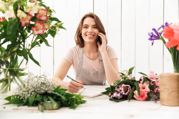 Young cute happy florist lady over flowers in workshop talking by mobile phone. - Φωτογραφία, εικόνα