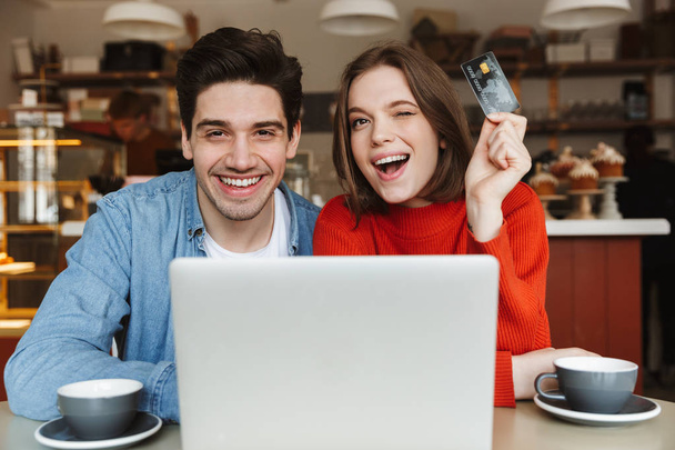 Excited young couple showing credit card and sitting at the cafe table with laptop computer - Photo, image