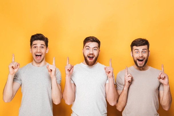 Three young happy men pointing up at copy space isolated over yellow background - Fotografie, Obrázek