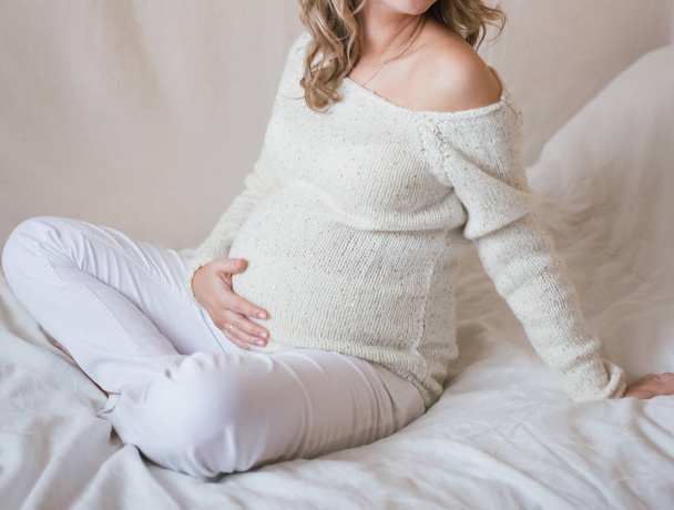 Беременная девушка сидит на кровати со скрещенными ногами и одной рукой гладит животик. Она одета в белые джинсы и вязаный белый обтягивающий свитер
. - Фото, изображение