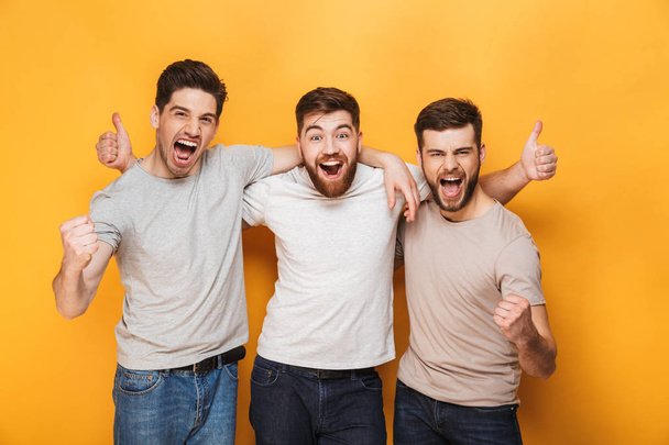 Three young excited men showing thumbs up and celebrating isolated over yellow background - Foto, afbeelding
