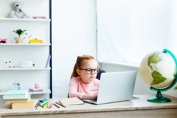 kaunis lapsi silmälaseissa kannettavan tietokoneen avulla opiskellessaan työpöydällä kotona
 - Valokuva, kuva