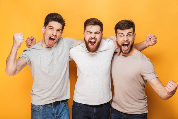 Three young excited men celebrating success together and screaming isolated over yellow background - Fotó, kép