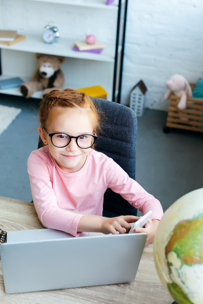 high angle view of adorable red haired schoolchild in eyeglasses smiling at camera while using smartphone and laptop - Fotó, kép