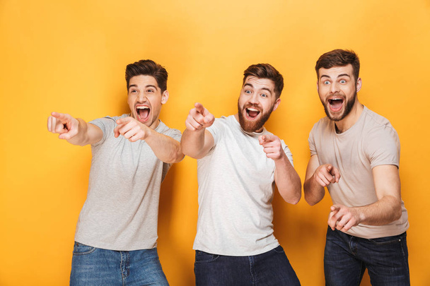 Three young excited men pointing away and screaming isolated over yellow background - Zdjęcie, obraz
