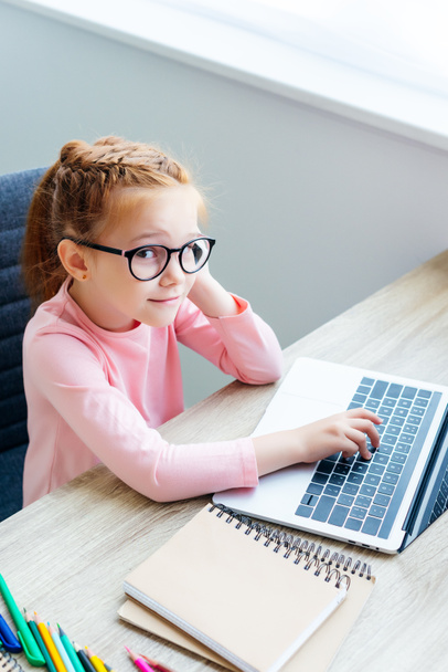 high angle view of smiling schoolchild i eyeglasses using laptop and looking at camera - Zdjęcie, obraz