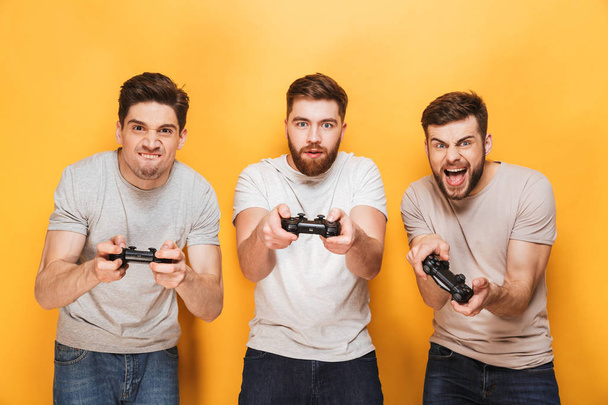 drei junge aufgeregte Männer, die Steuerknüppel in der Hand halten, während sie auf gelbem Hintergrund vereinzelt Videospiele spielen - Foto, Bild