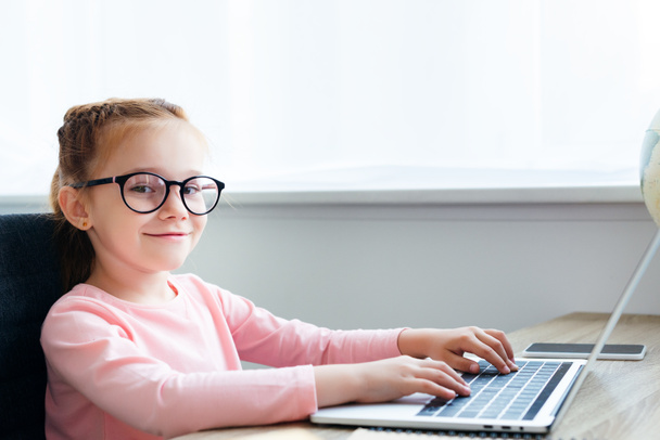 beautiful kid in eyeglasses using laptop and smiling at camera  - Foto, afbeelding