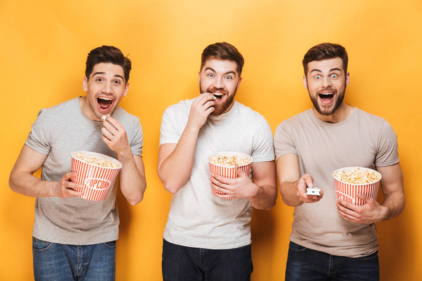 Three young excited men eating popcorn and looking at camera isolated over yellow background - Foto, Imagem