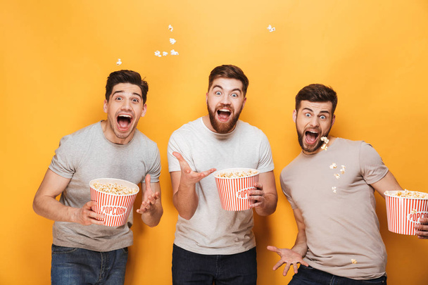 Three young excited men eating popcorn and celebrating isolated over yellow background - Photo, image