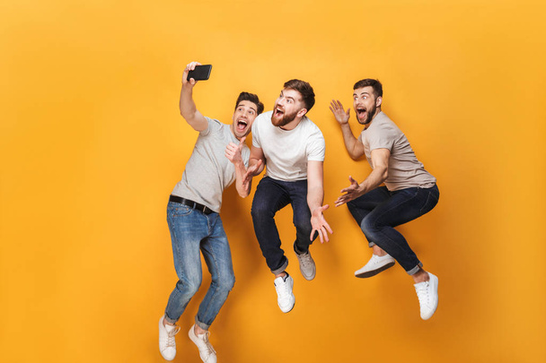 Three young smiling men taking a selfie together while jumping isolated over yellow background - Photo, image