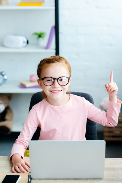 adorable red haired schoolgirl in eyeglasses pointing up with finger and smiling at camera while using laptop at home - Fotografie, Obrázek