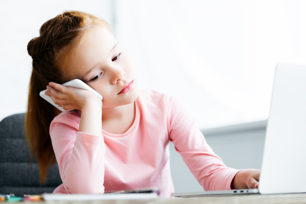 tylsistynyt pieni lapsi puhuu älypuhelimella ja käyttää kannettavaa tietokonetta kotona
 - Valokuva, kuva