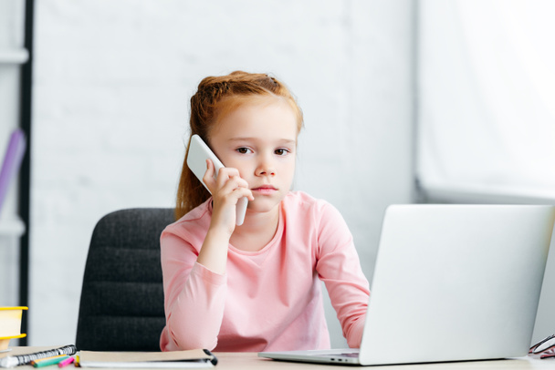 kaunis punapää lapsi puhuu älypuhelimella ja katselee kameraa kannettavan tietokoneen käytön aikana
  - Valokuva, kuva