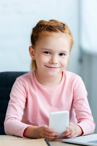 όμορφη κοκκινομάλλα παιδί χρησιμοποιώντας smartphone και να χαμογελά στη φωτογραφική μηχανή - Φωτογραφία, εικόνα